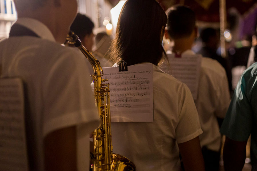 Um grupo de jovens, todos de camisa branca, caminham enfileirados pela rua à noite tocando instrumentos musicais. As partituras que leem, estão presas à camisa do músico à sua frente na fila.