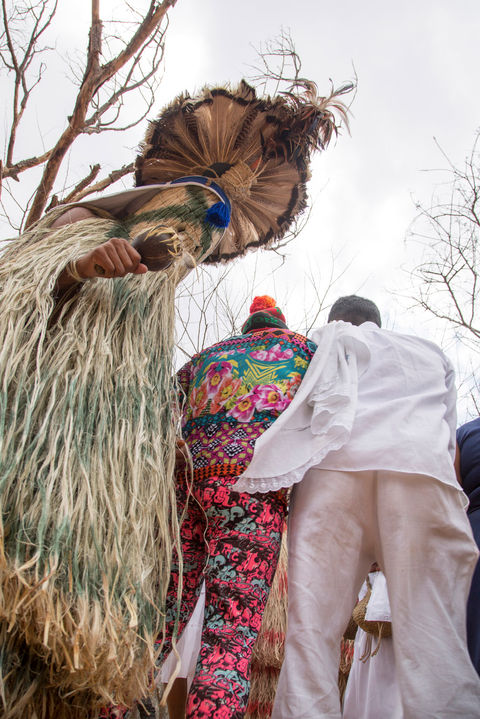 3 pessoas estão em pé, dançando. À esquerda, um pankararu com o traje do Toré - uma túnica de palha que cobre o corpo e o rosto, um chapéu redondo de palha e um chocalho na mão direita. Ao centro, de costas, uma pessoa com calça e blusa supercoloridas está abraçada a uma pessoa de calça e blusa brancas.