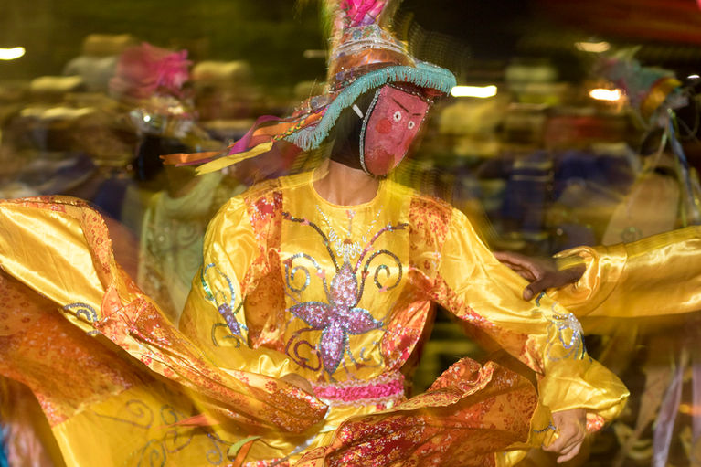 Um mascarado de vestido amarelo e chapéu de abas largas dança de forma vigorosa. A imagem, feita com baixa velocidade, capta os movimentos da saia e do corpo do brincante.