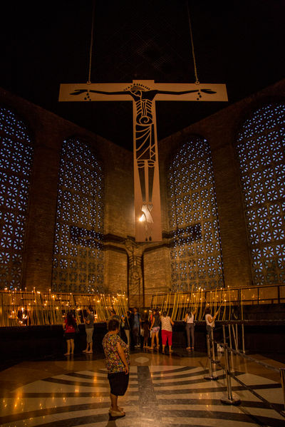 Na sala das velas, uma enorme cruz de Cristo, com design moderno domina o espaço. A sombra da cruz se projeta sobre a parede oposta. Embaixo dela, uma multidão acende suas próprias velas e ora.