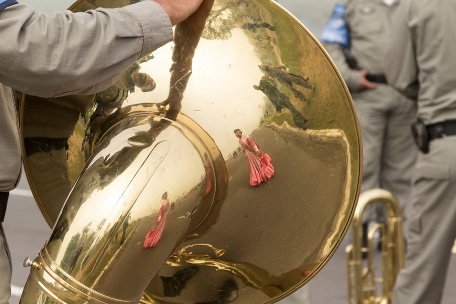Uma grande tuba dourada ocupa quase todo o espaço da foto. Vê-se que ela está sendo segurada por um homem de uniforme cinza, do qual só se enxerga o braço. Refletida no metal do instrumento, vem uma moça, caminhando pela rua, com um vestido longo alaranjado, a roupa típica das prendas gaúchas.