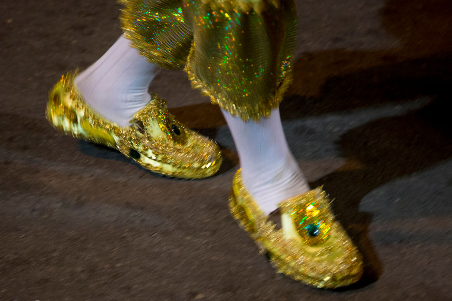 Os pés de um brincante de maracatu dançam. Ele usa um sapato todo enfeitado com lantejoulas e materiais dourados brilhantes. Usa meias brancas e uma calça meio curta, no mesmo tom dos sapatos.