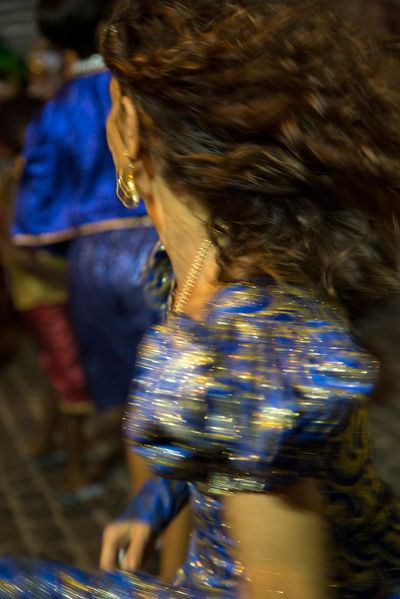 Uma mulher com vestido azul e dourado dança de forma animada. Não se vê o rosto da mulher, apenas o movimento do seu corpo e seu cabelo esvoaçante, captado com baixa velocidade na câmera.