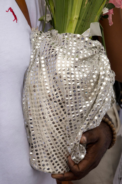 Um homem negro de camiseta branca segura um vaso de flores recoberto por um tecido prateado.