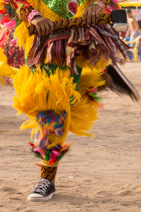 Close nos pés de uma cabocla de pena que está dançando vigorosamente. Suas mãos seguram a machadinha com força e seus movimentos denotam muita energia. As franjas amarelas de enfeites de perna se agitam pelo movimento da dança.