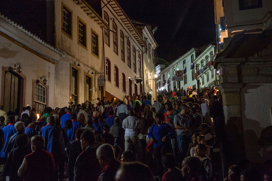 Na noite escura da Sexta-Feira Santa, uma multidão de fiéis caminha em procissão por uma rua de Ouro Preto. Ao fundo, de ambos os lados, é possível ver o casario colonial típico do centro histórico dessa cidade.