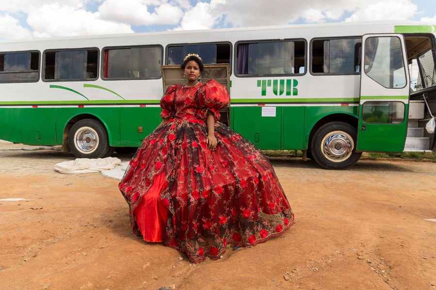 Uma moça negra está em pé sobre um chão de terra batida, em frente a um ônibus de turismo branco e verde. Ela está com uma pose muito altiva e usa um vestido de princesa vermelho, com bordados prateados e uma coroa na cabeça.