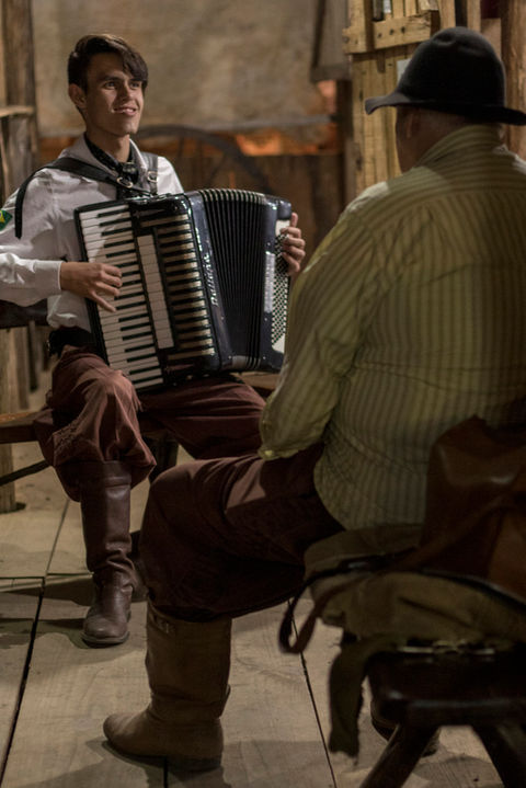 Dois homens estão sentados em uma varanda. O que está de frente para a câmera usa camisa branca, bombacha e bota marrom e está tocando um acordeão. O homem que está a frente dele e de costas para a imagem, usa uma camisa de manga comprida amarela, calça e bota marrom e chapéu preto.