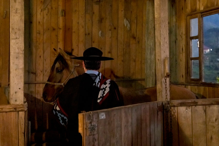 Um homem de poncho de lã e chapéu de feltro, ambos pretos, está de costas, dentro do estábulo, selando o seu cavalo. As paredes do estábulo são de madeira e, pela janela lateral da casa, à direita da imagem, nota-se que o dia está clareando lá fora.