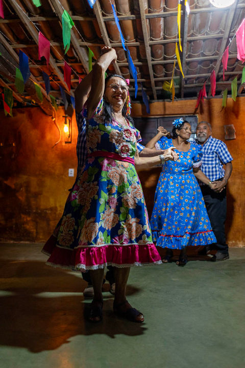 Em um salão bem escuro, várias pessoas dançam quadrilha. O salão tem o teto baixo, enfeitado com bandeirolas juninas. No lado esquerdo da imagem, em primeiro plano, uma senhora de vestido estampado rosa, azul e verde sorri enquanto dança com seu parceiro que está escondido atrás dela na imagem. Do lado direito, ao fundo, ve-se um oputro casal dançando a quadrilha. Ela de vestido azul com pequenas flores amarelas e ele de camisa quadriculada azul e branco e calça jeans. A parede ao fundo tem um tom de terra batida que deixa o ambiente bastante alegre.