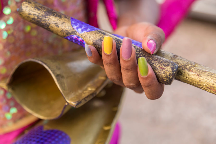 Destaque para as unhas da tocadora de agogô que segura as suas baquetas. Suas unhas são bem longas e arredondadas e estão pintadas cada uma de uma cor: lilás, amarelo, rosa antigo, verde e pink.