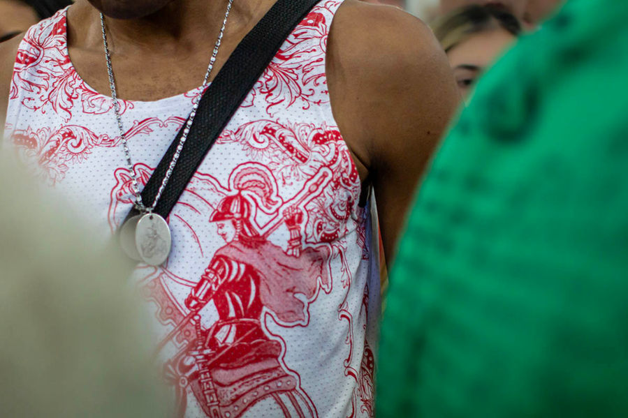 Destaque para a camiseta branca, com desenho vermelho de são Jorge sobre o cavalo. São Jorge empunha uma lança, num movimento de ataque. Não se vê o rosto do homem que usa a camiseta, apenas a sua estampa.