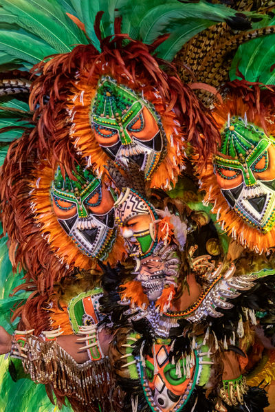 Um homem com uma fantasia super elaborada, cheia de máscaras, está em pé. Apesar da imagem enfocar o seu rosto, o excesso de máscaras na fantasia torna difícil a identificação da pessoa sob a fantasia.