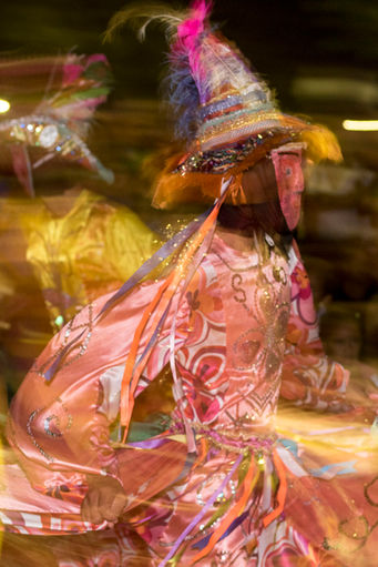 Um mascarado de vestido cor de rosa e chapéu de abas largas dança de forma vigorosa. A imagem, feita com baixa velocidade, capta os movimentos da saia do brincante.