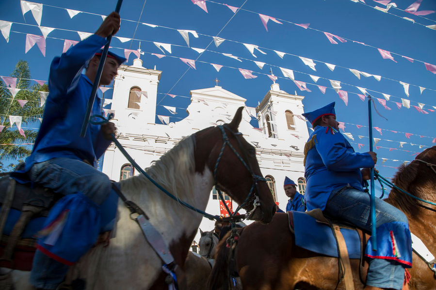 Dois homens montados em seus cavalos estão passando em frente à igreja matriz de São Luiz do Paraitinga, que pode ser vista ao fundo, contrastando com o céu completamente azul de um dia de muito sol.