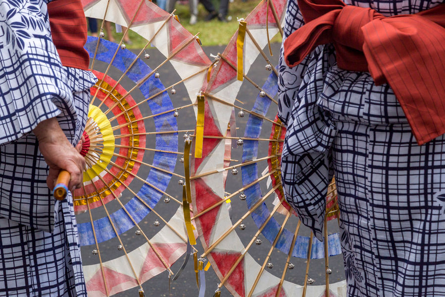 Os corpos de duas mulheres aparecem parcialmente na imagem. Cada uma delas segura uma espécie de guarda-chuva de papel colorido. As mulheres usam quimonos quadriculados em branco e preto e largas faixas vermelhas amarradas à cintura, à moda das gueixas.