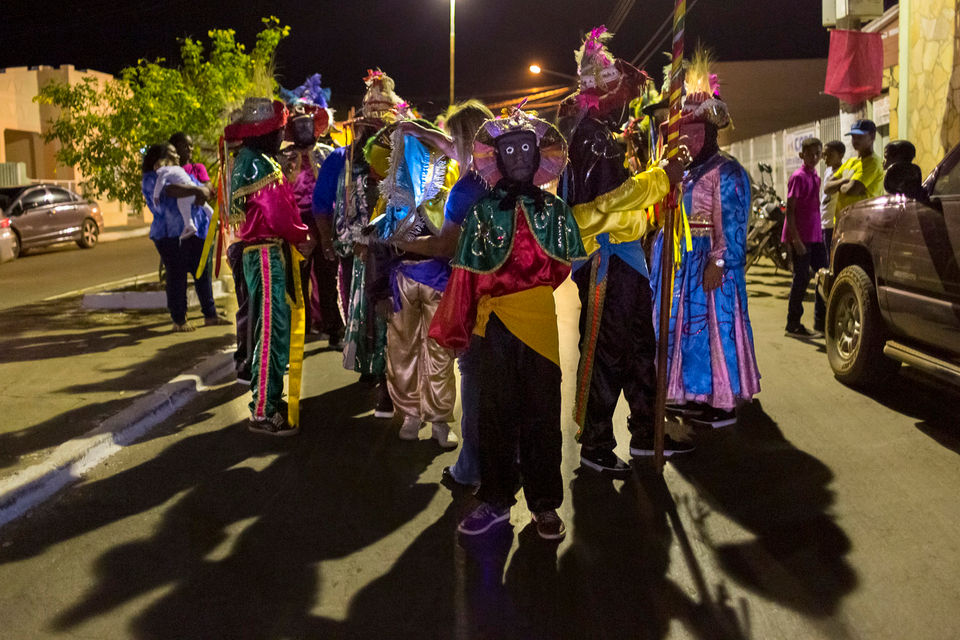 Um grupo de mascarados estão em pé no meio de uma rua aguardando o início da apresentação. Em volta deles, vê-se o movimento normal da cidade, com pessoas não fantasiadas e carros estacionados. 