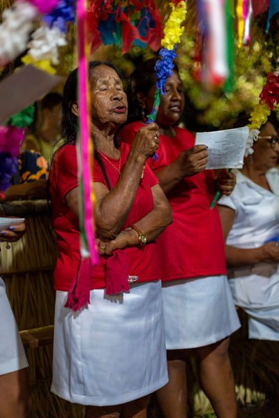 Uma senhora de camiseta vermelha e saia branca canta de olhos fechados enquanto segura uma bandeirola colorida.