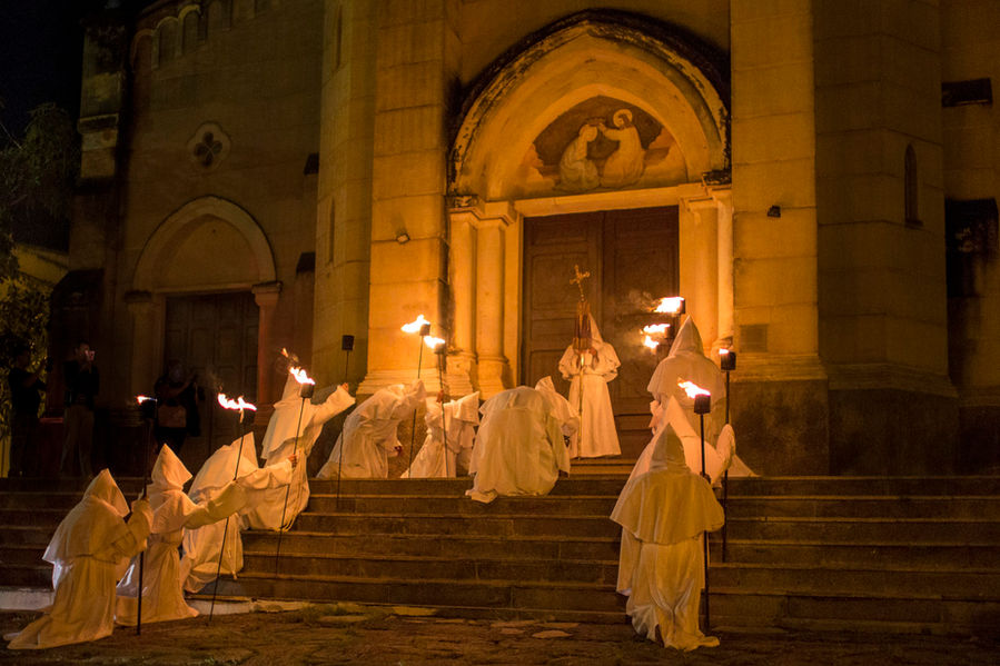 Um grupo de cerca de 12 pessoas, todas com túnicas longas e capuzes brancos, se enfileira nas escadarias de uma igreja. Cada uma dessas pessoas carrega uma tocha com fogo na ponta. Eles estão ajoelhados nos degraus, olhando para baixo, em posição de reverência, provavelmente rezando.