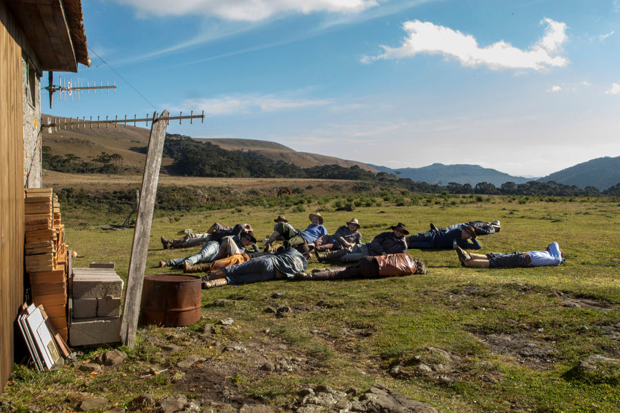 Em um grande gramado verdinho, cerca de 12 homens estão deitados, tomando sol. Eles estão todos vestidos com bombachas, botas, camisas de mangas longas e chapéus.