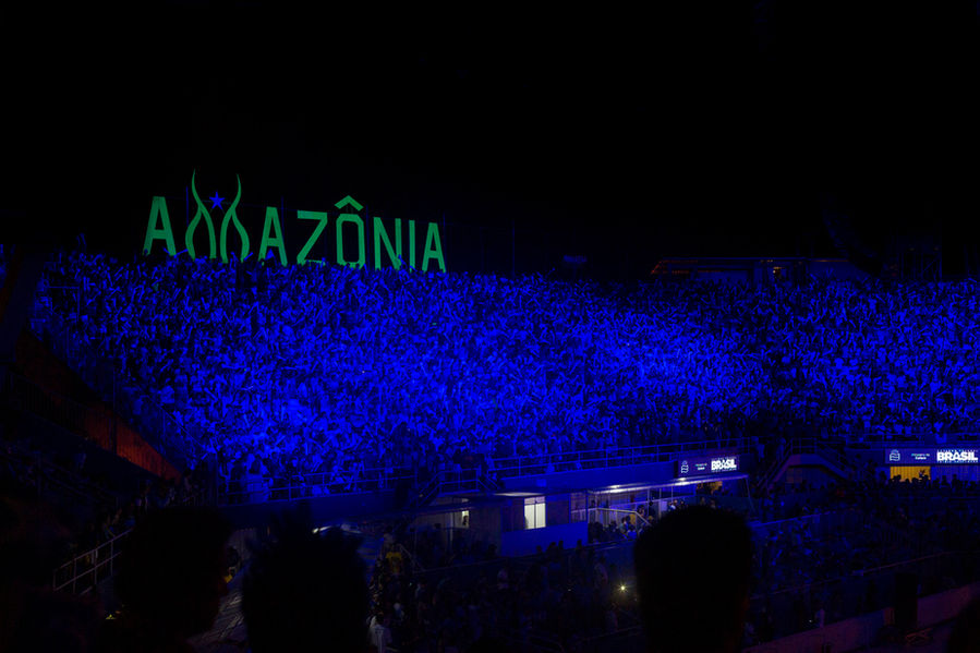 Vista panorâmica das arquibancadas do Bumbódromo onde estava a galera do boi Caprichoso. A iluminação faz a arquibancada parecer um mar bem azul e, mais acima, vê-se um letreiro com a palavra Amazônia escrita em verde.