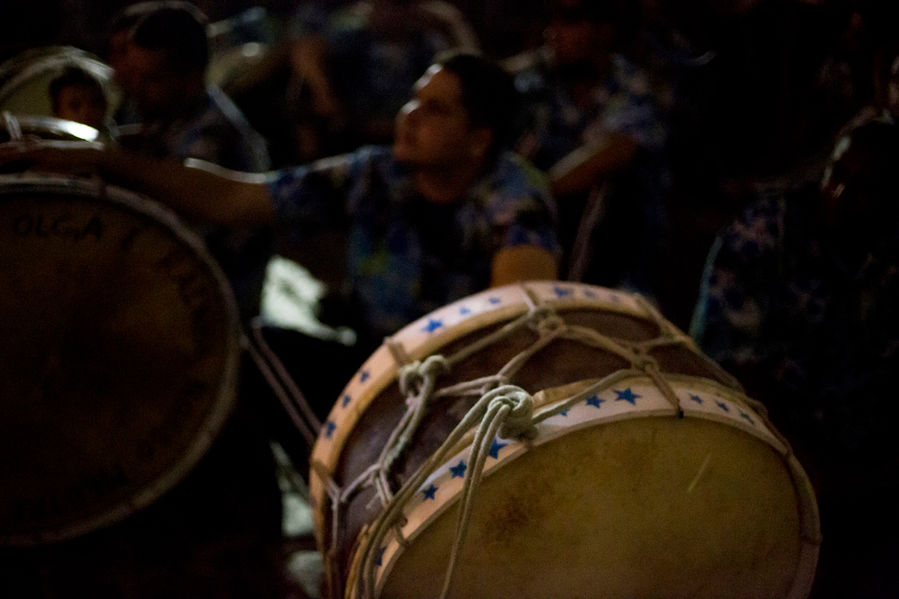 Numa noite bem escura, vários tambores repousam no chão. Alguns batuqueiros aparecem sentados próximos aos seus tambores, mas sem tocá-los.