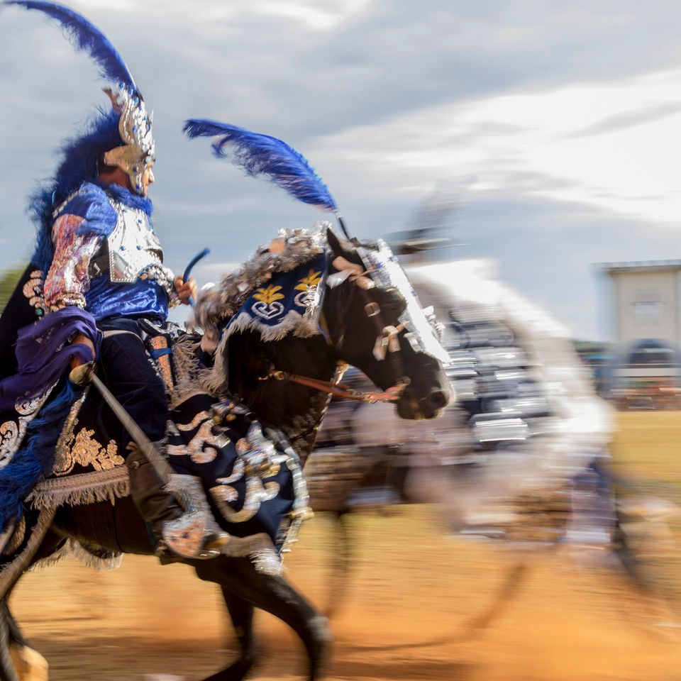 Dois homens e seus cavalos, adornados com roupas azuis ricamente bordadas em prateado, cavalgam em alta velocidade por um campo gramado. A imagem retrata o exato momento em que um cruzou o caminho do outro.