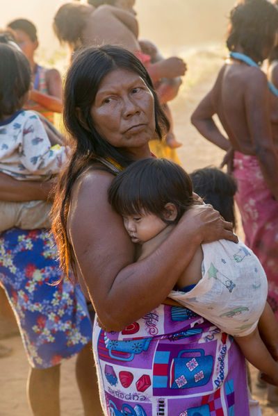 Uma mulher indígena com um pano colorido amarrado na cintura carrega um bebê adormecido. Atrás dela, menos nítidas, várias outras pessoas fazem a mesma coisa.