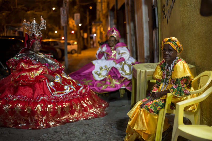 3 senhoras negras estão sentadas em cadeiras de plástico amarelo, na calçada em frente a um bar. Elas estão com fantasia de baiana. A da direita, com um vestido e turbante amarelos. A do centro, sentada mais ao fundo, usa um vestido rosa, branco e dourado e uma coroa na cabeça. A da esquerda, usa um vestido rodado gigante, vermelho e tem um candelabro de cristal na cabeça.