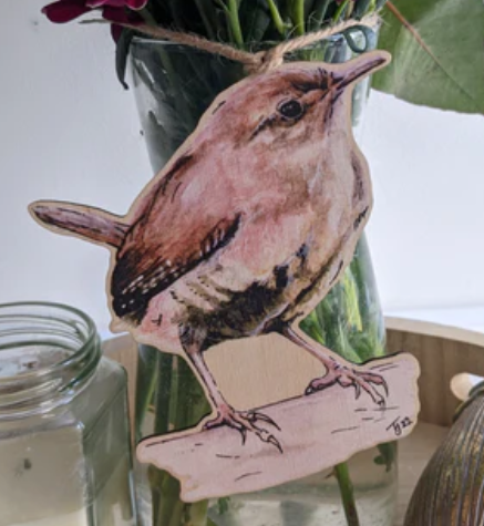 Wooden hanging decoration of a bird displayed on the front of a vase with flowers. A candle is nearby. Natural, earthy tones create a calm atmosphere.