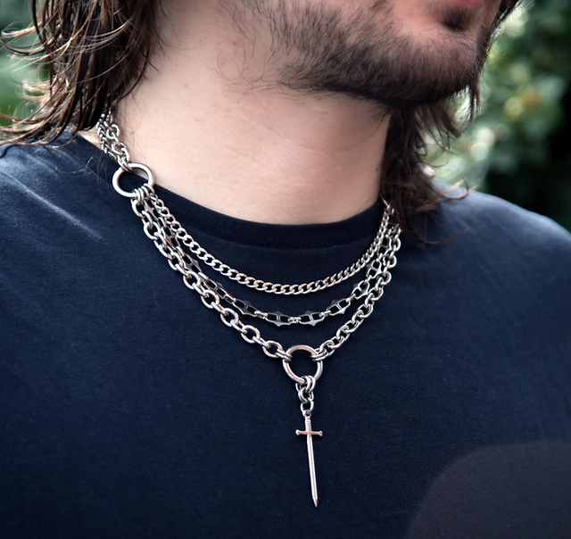 A bearded man wears a plain black t-shirt and a layered chain necklace featuring O-rings and a sword pendant.
