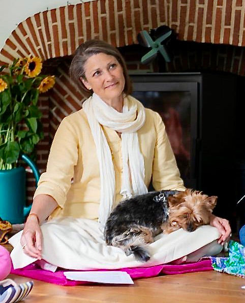 Picture of the yoga teacher, Ayurveda and mindfullness expert Susan Hopkinson with her dog, Meta in front of a fire place.
