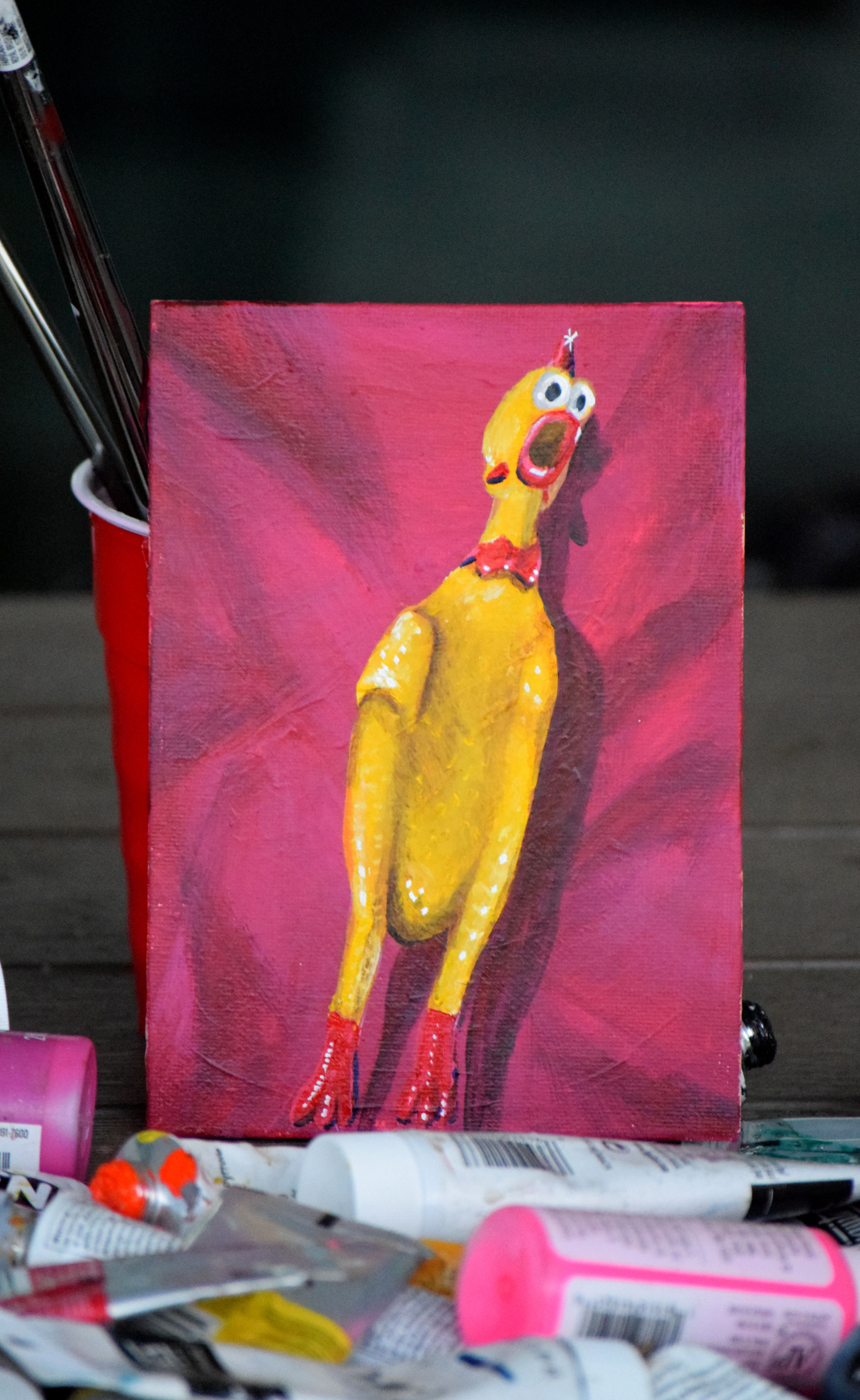Acrylic painting of a screaming yellow rubber chicken surrounded by art supplies.