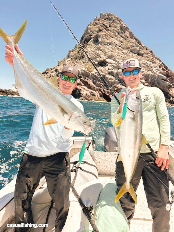 The photo above captures exactly that moment — two anglers standing on a center-console boat, deep blue water behind them, and a dramatic rocky outcrop rising straight from the sea. This is classic yellowtail country.