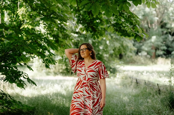 Woman in a red-and-white patterned dress stands under leafy trees in a sunny park, hand on head, looking thoughtful.