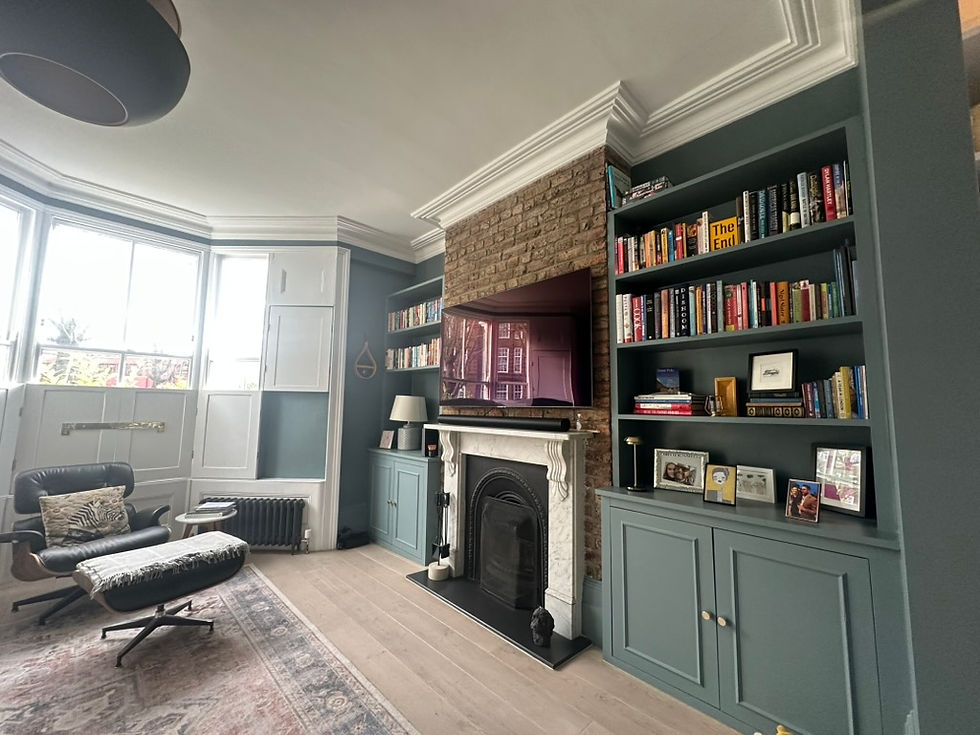 Cosy living room with a green bookshelf filled with books, a TV above a brick fireplace, an armchair, and a large window.
