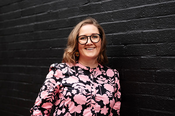 A photograph of Hannah Ashe wearing a pink and black floral dress standing in front of a black wall