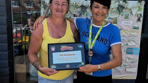 Two women are smiling to the camera, one has her arm around the other. One woman in a yellow vest is holding a plaque.