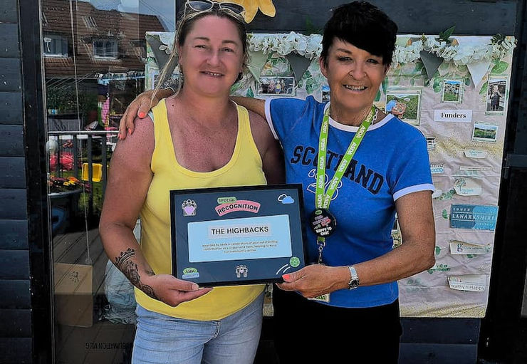 Two women are smiling to the camera, one has her arm around the other. One woman in a yellow vest is holding a plaque.