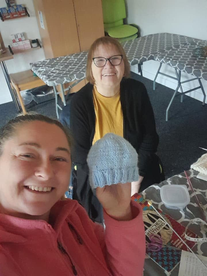 Two women are indoors and smiling to the camera. One holds up a knitted baby hat. Crafting supplies are on the table.