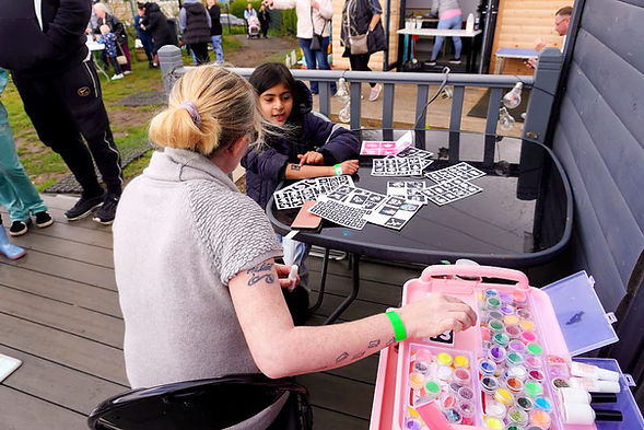 A volunteer applies a temporary glitter tattoo to a young girl at the High Backs Easter event in Burnhill, Rutherglen. Stencils and a colourful case of glitter are spread across the table on the main hall decking, with other visitors visible in the background.