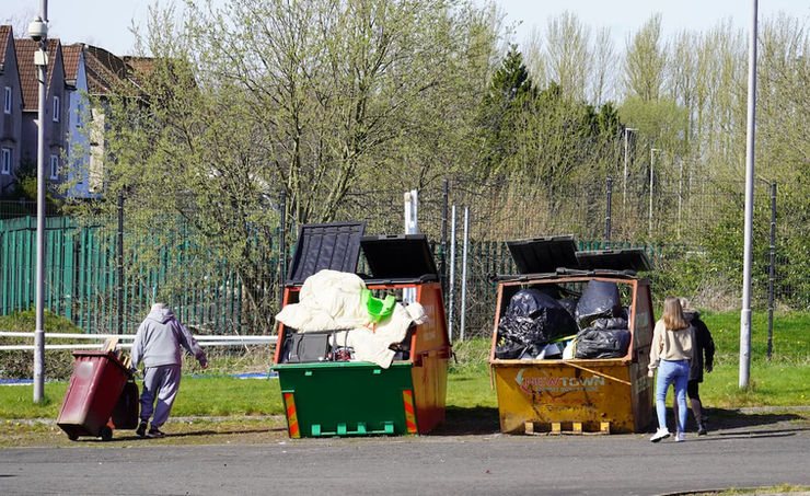 A man drags a wheelie bin towards two large skips which appear very full.