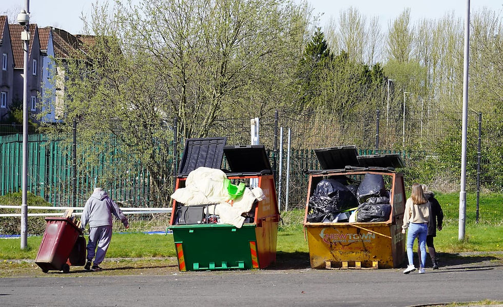 A man pulls a wheelie bin with timber towards two large skips which are very full. A woman and another person stand beside one of the skips.