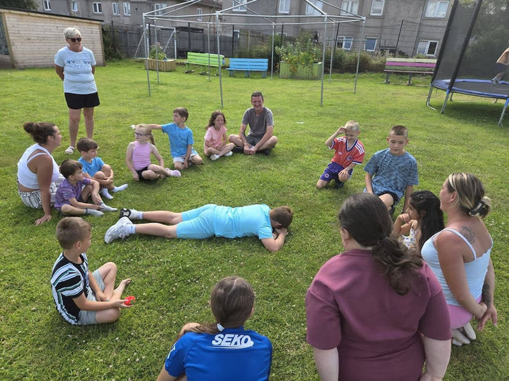 Children and three adults are seated in a circle on grass. One child lies face down in the center taking part in a game or wellbeing exercise.
