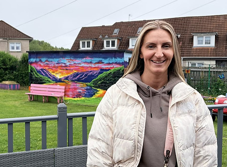 Julie-Ann stands in the High Backs community garden in front of a colourful mural of a mural of a Scottish loch scene painted on the side of a shed. Houses on Penman Avenue, Burnhill are in the background.