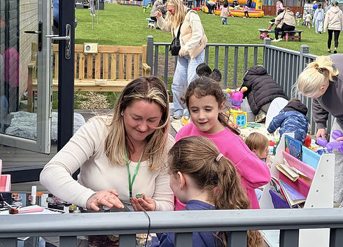 Lizzy McDonald, founder of The High Backs, doing craft activities with two children on the decking at The High Backs community garden in Burnhill, with families gathering in the garden behind them.