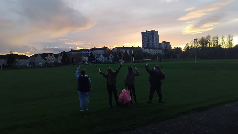 The High Backs kids celebrate in a playing field in Burnhill. They are holding a litter picking bag and sticks. The background sunset can be seen as they raise their arms in triumph.