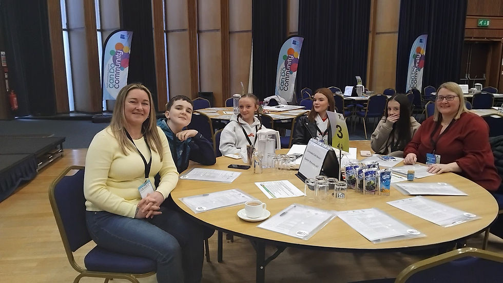 Two adults sat with four youths around a round table at an official event. Flags in the background say Can Do Community.