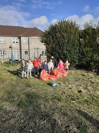 A group of children stand together on scrubby ground in Burnhill holding red litter bags and a football, smiling at the camera after a community litter pick.