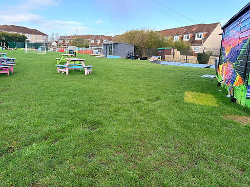 A broad expanse of green grass enclosed by residential houses. Brightly coloured picnic tables are scattered across the lawn. A large colourful mural is painted on the side of a storage unit to the right, with a community building visible further along.